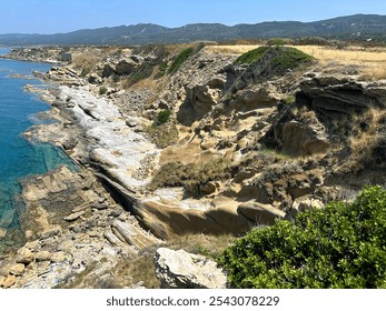 The rocky coastline features layered stone formations and lush green plants along the edge, under a clear blue sky and bright sunlight, creating a tranquil seascape. - Powered by Shutterstock