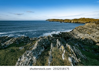 A rocky coastline with green vegetation is seen on a clear day with a blue sky. The ocean stretches out in front of the shore, and the waves are breaking on the rocks. - Powered by Shutterstock