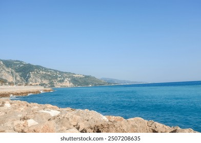 rocky coastline meets clear blue waters under a bright sky with distant hills stretching along the serene shoreline - Powered by Shutterstock