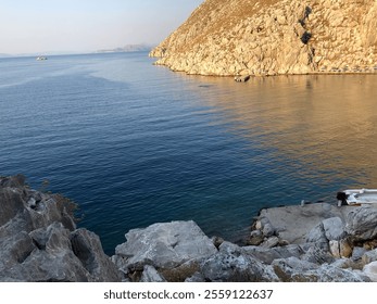 A rocky coastline overlooking calm, deep blue sea with rugged hills in the background under clear skies - Powered by Shutterstock