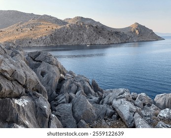 A rocky coastline overlooking calm, deep blue sea with rugged hills in the background under clear skies - Powered by Shutterstock