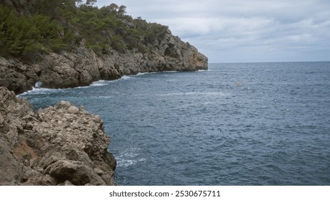 Rocky coastline with pine trees and calm blue waters in cala deia, mallorca under an overcast sky - Powered by Shutterstock