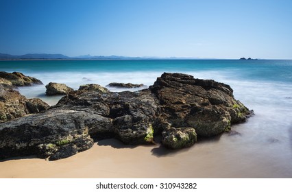 Rocky Coastline With Sand And Misty Ocean Below The Cape Byron Lighthouse And Overlooking the Julian Rocks Island with a Beautiful Clear Blue Sky, Wategos Beach, Byron Bay, New South Wales, Australia