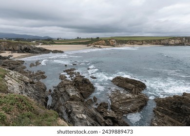 rocky coastline with waves crashing under a cloudy sky, surrounded by green fields and distant mountains - Powered by Shutterstock
