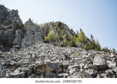 A rocky hillside with trees atop, displaying terrestrial plants and mountainous formations under the sky, presenting a natural landscape. Tall trees grow on the summit, forming a picturesque scene - Powered by Shutterstock