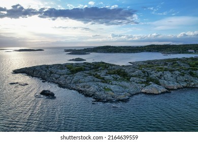 Rocky land with sea, sun setting and small islands around. Bits of green plants in the rocks. Two people standing on the rocky island.