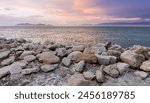  A rocky lined shoreline overlooking the Great Salt Lake in Utah with small silhouette mountain range in the background as the sun sets casting pastel colors of pink, blue, orange, and purple. 