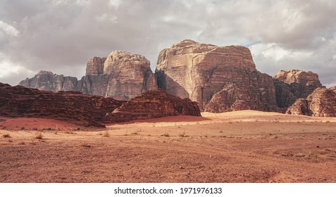 Rocky Massifs On Red Sand Desert, Few Dry Grass Cluster Ground, Cloudy Sky In Background, Typical Scenery In Wadi Rum, Jordan