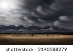 Rocky Mountain landscape scene during the winter with snow-capped peaks and a meadow in the foreground with a dramatic dark sky and wispy white clouds.