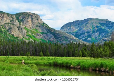 Rocky Mountain National Park lake in July with mountains and trees in the background and a lake, grass and a man fly fishing in the foreground - Powered by Shutterstock