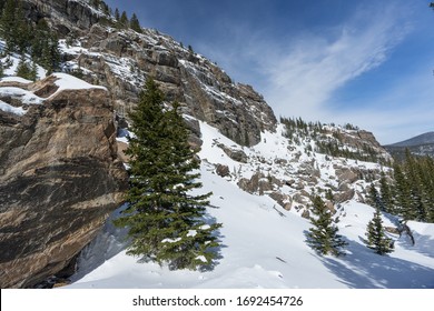 Rocky Mountain National Park Snowy Winter Landscape Of Rocky Cliff With Pine Trees And Blue Sky And Wispy Clouds