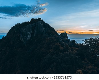 Rocky mountain peak with lush trees, silhouetted against a vibrant sunset sky with scattered clouds. - Powered by Shutterstock