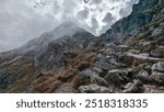 A rocky mountain trail winds upward through a rugged landscape, surrounded by mist and dramatic clouds. The steep path and rough terrain evoke a sense of challenge and adventure in the wilderness.