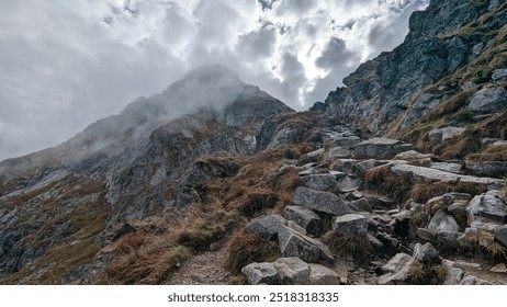 A rocky mountain trail winds upward through a rugged landscape, surrounded by mist and dramatic clouds. The steep path and rough terrain evoke a sense of challenge and adventure in the wilderness. - Powered by Shutterstock