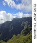 Rocky peaks of Pico Ruivo pierce through white clouds under a clear blue sky, surrounded by lush green slopes—Madeira