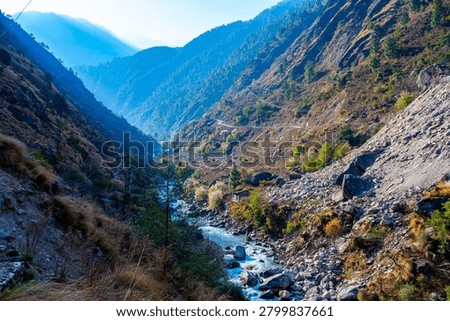 A rocky river flows through a deep mountain valley in Gosaikunda. Steep slopes covered in sparse vegetation and a small building are visible under a bright sky. Gosaikunda, Nepal