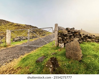 Rocky and rugged mountain trail leading uphill. A closed chain link fence run across the gravel road. A stone wall and grassy green hills surround the scene. Nólsoy, Faroe Islands. - Powered by Shutterstock