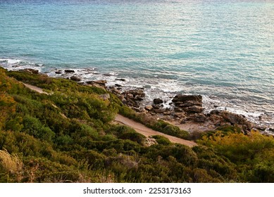 Rocky sea coast with sea waves and green bushes on the slopes. Sand on the shore, rocky boulders in the sea. The natural landscape of the island of Rhodes