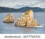 Rocky sea stack at a tranquil Basque coastline - A long exposure of a large rock formation in the middle of the sea with smooth water and rolling mountains in the background.