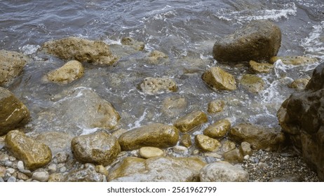 Rocky seashore with wet stones and gentle waves crashing under cloudy skies suggesting a serene coastal landscape. - Powered by Shutterstock