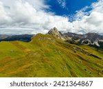 Rocky Seceda ridgeline overgrown with green grass on sunny day. Light clouds and giant forestry mountains around valley in Italian Alps aerial view