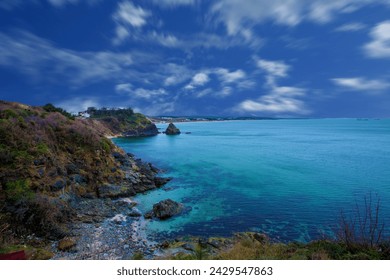 A rocky shore with a body of water and a blue sky with clouds, a body of water surrounded by a lush green hillside 