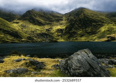 A rocky shore slopes down to a still, dark water lake, with two large, green mountains in the distance. - Powered by Shutterstock