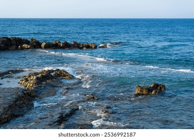 A rocky shoreline with gentle waves washing over the stones, set against the vast, tranquil blue sea under a clear sky. Blue sea. Shoreline. Waves. - Powered by Shutterstock