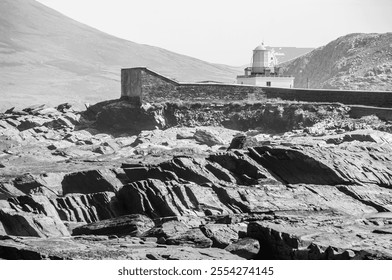 A rocky shoreline with a lighthouse in the distance. The lighthouse is a small building with a white roof. The scene is peaceful and serene - Powered by Shutterstock