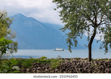 Rocky shoreline with lush vegetation in idyllic lakeside town Nago-Torbole, Lake Garda, Trentino, Italy. ferry boat is sailing on the calm waters surrounded by steep rugged mountains of Garda Prealps - Powered by Shutterstock