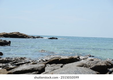 A rocky shoreline meets the clear blue sea, with gentle waves lapping against the stones. The serene coastal landscape invites a sense of calm and relaxation - Powered by Shutterstock