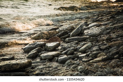 A rocky shoreline on the banks of Lake Ontario shimmers in the evening twilight. Small cresting waves flow toward large wet stones at water’s edge. Warm shades of gray, yellow and pale blue. - Powered by Shutterstock