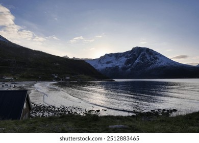 A rocky shoreline with scattered snow and sparse trees, overlooking a peaceful lake and distant mountains under a soft cloud-filled sky at sunset. - Powered by Shutterstock