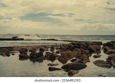 rocky shoreline with waves crashing against the tidal pool wall  under a cloudy sky. The water is relatively calm near the shore, reflecting the rocks and the cloudy sky. - Powered by Shutterstock