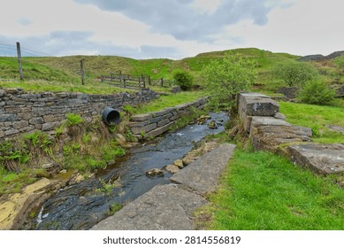 A rocky stream flows between old stone walls at Cheesden Lumb Mill, Bury, UK, with a black pipe emptying into it.