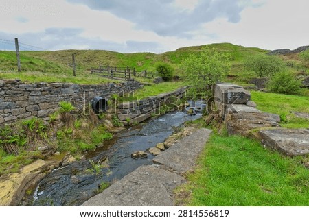 A rocky stream flows between old stone walls at Cheesden Lumb Mill, Bury, UK, with a black pipe emptying into it.