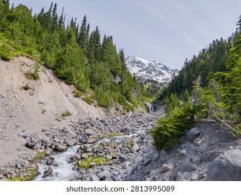 A rocky stream flows through a forested valley with a waterfall and a snow-capped mountain peak in the background.