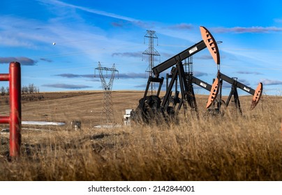 Rocky View County Alberta Canada, March 28 2022: Oil Pump Jacks Working In An Agriculture Field With Distant Electrical Power Lines.