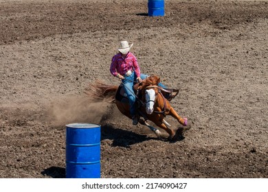 A rodeo cowgirl in a red shirt is riding a brown horse in a barrel racing competition. They are going around the barrel on the left side.  The horse is kicking up a lot of dirt.