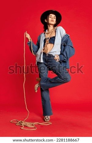 Rodeo. Portrait of beautiful young woman in jeans clothes and black hat posing with string like cowboy against red studio background. Concept of style, beauty, fashion, youth, emotions. Ad