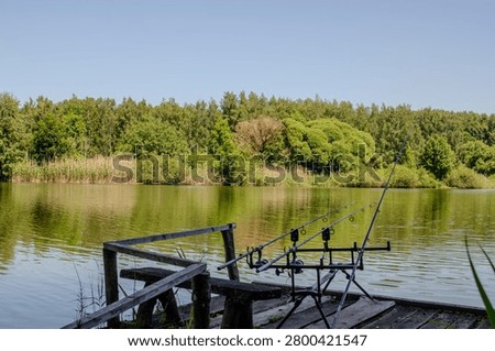 ​Fishing rods setup on a rod pod holder on a wooden pier by a calm summer lake.