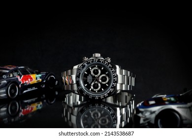 Rolex wristwatch model cosmograph daytona oyster perpetual superlative chronometer with black ceramic bezel stainless steel body on black table with sports racing cars models at authorized dealer shop