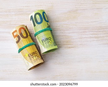 Roll Of 100 Euro Banknotes And Wad Of 50 Euro Paper Currency On A White Wooden Desk. Cash Savings, Counting Money, Income And Expenses Concepts. Copy Space. Top View.