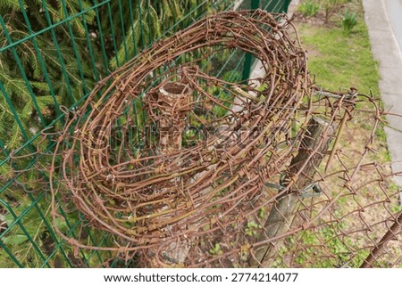 Roll of the old rusted single strand barbed wire lies on a metal pole on a blurred background of a modern metal lattice fence at overcast day