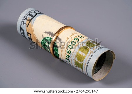 A rolled 100 US dollar banknote featuring Benjamin Franklin, held by a gold ring on a grey surface.