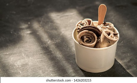 Rolled ice cream with in cone cup on dark rustic background. Iced rolls with biscuits chocolate sandwich cookies. Thai style rolled ice cream with copy space for text or design. Banner. Hard light