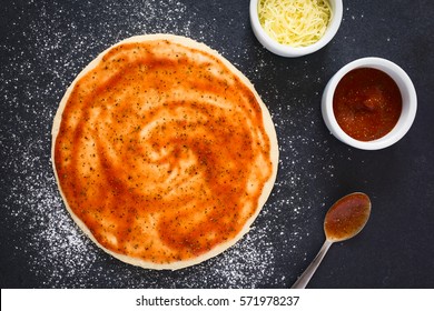 Rolled Out Pizza Dough With Tomato Sauce On Floured Slate Surface, Tomato Sauce And Grated Cheese On The Side, Photographed Overhead With Natural Light