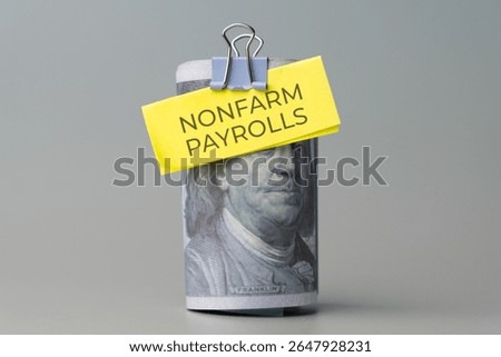 A rolled stack of US one-hundred dollar bills with a note "NONFARM PAYROLLS" attached. Economic indicator, labor market data, employment statistics concept.