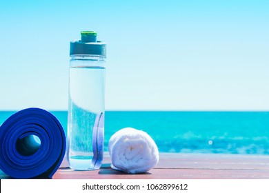 Rolled Yoga Mat Bottle with Water White Towel on Beach with Turquoise Sea Blue Sky in Background. Sunlight. Relaxation Summer Meditation Fitness Wellbeing Mindful Living Concept. Copy Space