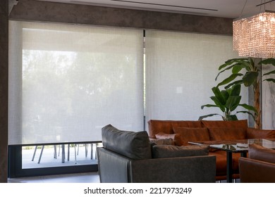 Roller Blinds In The Interior. Automatic Solar Shades Large Size On The Window. Living Room Interior With Sofas And Palm Trees. Electric Sunscreen Curtains For Home. 